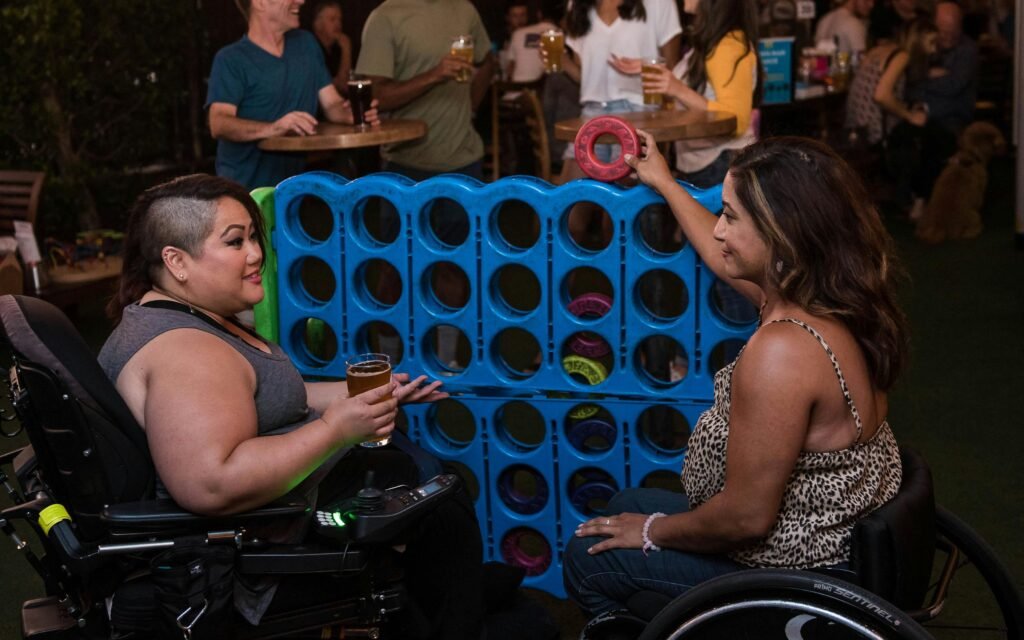 Two women in wheelchairs playing connect four at an indoor bar setting, enjoying a lively social atmosphere.